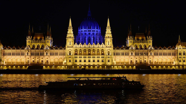 En la imagen se observa un barco turístico navegando por el Danubio frente al Parlamento húngaro completamente iluminado de azul. 